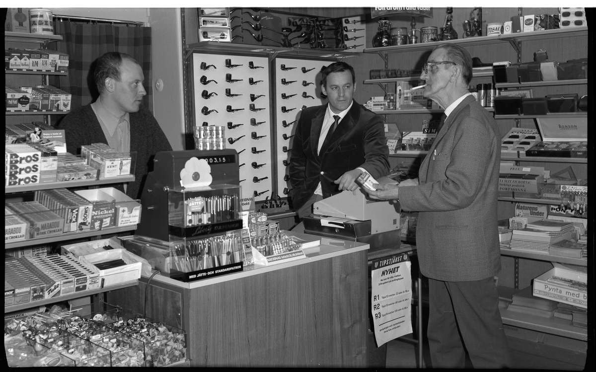 Cigarrboden på Göteborgsvägen 1, i kv Cederberg.
OBS aktuell adress vid fotograferingstillfäller, idag Magasinsgatan.
Åke Matsson och Gert Andersson.

Bakom disken i tobaksbutiken står två män.
På en stor tavla bakom dem (till höger om en öppning i vilken det hänger ett rutigt draperi) är pipor monterade, sex rader med ca 10 pipor i varje. 

På disken står en kassaapparat, ett ställ med batterier och ett ställ med pennor.
På kassaapparaten sitter en stor majblomma.

På en rullvagna står en särskild apparat som det står Tipstjänst på.

På en hylla till vänster om öppningen står bl a cigarettpaket, på andra hyllor som täcker väggarna finns blandade produkter.

Framför disken står en äldre man med ett cigarettpaket, Prince, i handen och en cigarett i munnen.



Foto 12.5.1969