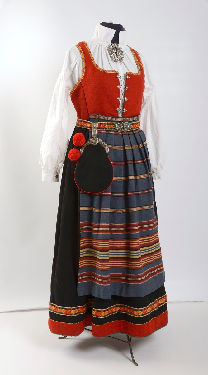 Kvinnebunad fra Vestfold, 1956 - modell.

Giver og tidligere eier, Wenche Blomberg, har fortalt dette om bunaden: 
"Vestfoldbunaden omtales i Norsk bunadleksikon b. 2, min er 1956-modellen avbildet på s.99.Jeg kjøpte den brukt på 1970-tallet en gang. Selgeren hadde sydd den selv og initialene hennes står på et lite metallmerke som er festet på innsida av capen. Husker ikke navnet. Hun bodde på Nøtterøy-sida av Kanalbrua i Tønsberg. Den passet akkurat, så jeg slapp å bruke penger på tilpassing. Jeg bare sydde på et par ekstra smålommer på den grønne understakken etter en tid. Bortsett fra snøringslenka som fulgte med, kjøpte jeg det øvrige sølvet sjøl. Lue og sko likeså. Etter noen år med heftig dansing der lua hele tida føyk av fordi jeg ikke likte å bruke hakeknyting, sydde jeg på en borrelås-strimmel inni, og da satt den bedre fast i håret. Ekstra bomullsskjorte kjøpte jeg seinere på Husfliden i Tønsberg.
 
Brukte den lite de første åra, men da jeg kom tilbake til Oslo i 1985 etter fem år i Nord-Gudbrandsdalen og etterhvert ble leikarring-aktiv, økte bruken betraktelig. I skjørtet er det ei lita rift som jeg rev opp da jeg klatra over et nettinggjerde i hagen utafor bygningen som tidligere huset Norges geografiske oppmåling, seinere Kunstakademiet, der det ofte var forskjellige festlige arrangementer. Jeg husker ikke anledningen og hvorfor jeg ble hengende fast i det gjerdet, men skaden var ikke større enn at jeg reparerte den sjøl. Pompongene på forklesnora var ellers et sårt punkt, måtte få noen til å lage nye for meg en gang, men jeg husker ikke hvem som gjorde det - kanskje Husfliden ett eller annet sted. Låsen på lauslomma var også til stadig irritasjon fordi den sprang opp hele tida så tobakken min datt ut.

Når man er folkedanser er skoa nærmest hjertet! Disse skoa var til slutt så godt inntrødde at jeg kvidde meg for å bruke et par nye jeg kjøpte via BUL. (Forresten: Hvilket par fikk dere? De gamle slitte, eller de nye? Begge?) [Opplysning fra Kvinnemuseet: Bare de nye skoene er del av gaven]

Seinere på 80-90-tallet en gang fikk jeg lagt stakken ut i livet av systova på Heimen. De bildene jeg har funnet er fra tida i Leikarringen i BUL. Det er forresten ikke lett å ri med bunadsstakk, kan jeg meddele. Hvor skal man gjøre av alt stakkestoffet der man sitter overskrevs? 

Jeg brukte bunaden siste gang 17. mai 2017 i Svullrya. Da hadde den krympa så mye at jeg knapt fikk den på meg, og nå så jeg ingen grunn til å bruke mer penger på utvidelser."

Til bunaden hører også to jakkemerker, ett fra Noregs ungdomslag, NU, og ett med symbol av en drage, som er merket for mikrostaten Ladonia, der Wenche Blomberg er Minister of Madness og Enkegrevinne Wenche von Blomberg und Grünerløkka. 

Om det siste merket forteller Wenche Blomberg: 
"Ladonia: Virtuell ministat opprettet av kunstneren Lars Vilks. Det finnes en god del slike på nettet, men Ladonia er i tillegg knyttet til et konkret geografisk område ved Kullen i Skåne: ei nesten utilgjengelig strandstripe av kjemperullestein der Vilks har bygd opp en rekke monumentale skulpturer av rekved. Historie, bilder etc på www.ladonia.org. Det har forekommet åratals med fornøyelige tvister med grunneiere og staten Sverige. Alle kan bli borgere av Ladonia, noe som fra tid til annen har ført til massehenvendelser fra en del land i Asia fra folk som vil emigrere og søker visum. Statsbudsjettets inntektsside stammer i stor grad fra salg av titler. Det var sånn jeg ble enkegrevinne og står i adelskalenderen. Man kan foreslå seg selv til minister for et hvilket som helst fantasirikt fagområde, men dette må godkjennes av regjeringen. Det var ingen innvendinger mot min søknad om å bli «minister of madnesss", man fant at den passet utmerket inn."