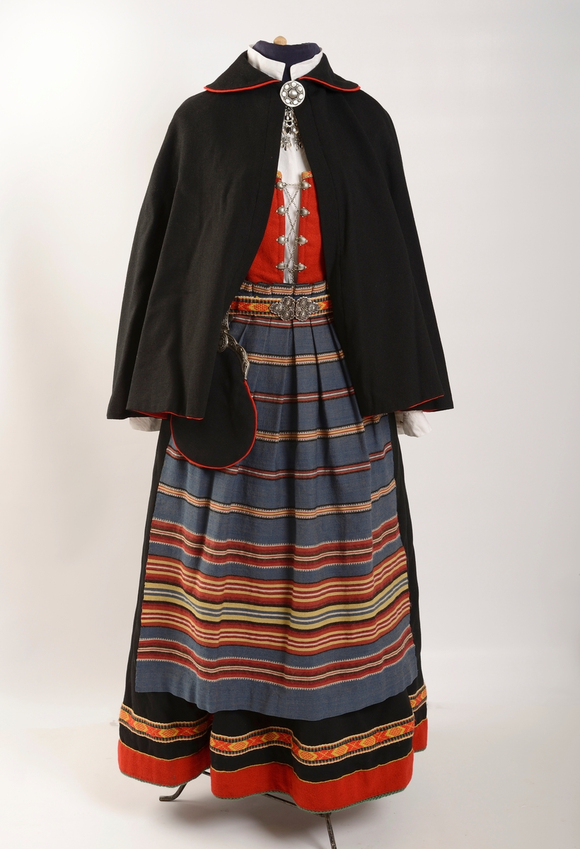 Kvinnebunad fra Vestfold, 1956 - modell.

Giver og tidligere eier, Wenche Blomberg, har fortalt dette om bunaden: 
"Vestfoldbunaden omtales i Norsk bunadleksikon b. 2, min er 1956-modellen avbildet på s.99.Jeg kjøpte den brukt på 1970-tallet en gang. Selgeren hadde sydd den selv og initialene hennes står på et lite metallmerke som er festet på innsida av capen. Husker ikke navnet. Hun bodde på Nøtterøy-sida av Kanalbrua i Tønsberg. Den passet akkurat, så jeg slapp å bruke penger på tilpassing. Jeg bare sydde på et par ekstra smålommer på den grønne understakken etter en tid. Bortsett fra snøringslenka som fulgte med, kjøpte jeg det øvrige sølvet sjøl. Lue og sko likeså. Etter noen år med heftig dansing der lua hele tida føyk av fordi jeg ikke likte å bruke hakeknyting, sydde jeg på en borrelås-strimmel inni, og da satt den bedre fast i håret. Ekstra bomullsskjorte kjøpte jeg seinere på Husfliden i Tønsberg.
 
Brukte den lite de første åra, men da jeg kom tilbake til Oslo i 1985 etter fem år i Nord-Gudbrandsdalen og etterhvert ble leikarring-aktiv, økte bruken betraktelig. I skjørtet er det ei lita rift som jeg rev opp da jeg klatra over et nettinggjerde i hagen utafor bygningen som tidligere huset Norges geografiske oppmåling, seinere Kunstakademiet, der det ofte var forskjellige festlige arrangementer. Jeg husker ikke anledningen og hvorfor jeg ble hengende fast i det gjerdet, men skaden var ikke større enn at jeg reparerte den sjøl. Pompongene på forklesnora var ellers et sårt punkt, måtte få noen til å lage nye for meg en gang, men jeg husker ikke hvem som gjorde det - kanskje Husfliden ett eller annet sted. Låsen på lauslomma var også til stadig irritasjon fordi den sprang opp hele tida så tobakken min datt ut.

Når man er folkedanser er skoa nærmest hjertet! Disse skoa var til slutt så godt inntrødde at jeg kvidde meg for å bruke et par nye jeg kjøpte via BUL. (Forresten: Hvilket par fikk dere? De gamle slitte, eller de nye? Begge?) [Opplysning fra Kvinnemuseet: Bare de nye skoene er del av gaven]

Seinere på 80-90-tallet en gang fikk jeg lagt stakken ut i livet av systova på Heimen. De bildene jeg har funnet er fra tida i Leikarringen i BUL. Det er forresten ikke lett å ri med bunadsstakk, kan jeg meddele. Hvor skal man gjøre av alt stakkestoffet der man sitter overskrevs? 

Jeg brukte bunaden siste gang 17. mai 2017 i Svullrya. Da hadde den krympa så mye at jeg knapt fikk den på meg, og nå så jeg ingen grunn til å bruke mer penger på utvidelser."

Til bunaden hører også to jakkemerker, ett fra Noregs ungdomslag, NU, og ett med symbol av en drage, som er merket for mikrostaten Ladonia, der Wenche Blomberg er Minister of Madness og Enkegrevinne Wenche von Blomberg und Grünerløkka. 

Om det siste merket forteller Wenche Blomberg: 
"Ladonia: Virtuell ministat opprettet av kunstneren Lars Vilks. Det finnes en god del slike på nettet, men Ladonia er i tillegg knyttet til et konkret geografisk område ved Kullen i Skåne: ei nesten utilgjengelig strandstripe av kjemperullestein der Vilks har bygd opp en rekke monumentale skulpturer av rekved. Historie, bilder etc på www.ladonia.org. Det har forekommet åratals med fornøyelige tvister med grunneiere og staten Sverige. Alle kan bli borgere av Ladonia, noe som fra tid til annen har ført til massehenvendelser fra en del land i Asia fra folk som vil emigrere og søker visum. Statsbudsjettets inntektsside stammer i stor grad fra salg av titler. Det var sånn jeg ble enkegrevinne og står i adelskalenderen. Man kan foreslå seg selv til minister for et hvilket som helst fantasirikt fagområde, men dette må godkjennes av regjeringen. Det var ingen innvendinger mot min søknad om å bli «minister of madnesss", man fant at den passet utmerket inn."