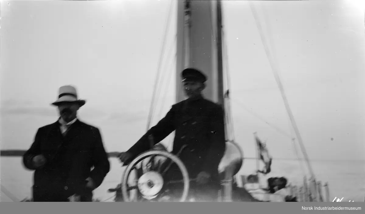 To menn på dekk på dampyachten Alexandra, en mann står ved roret.