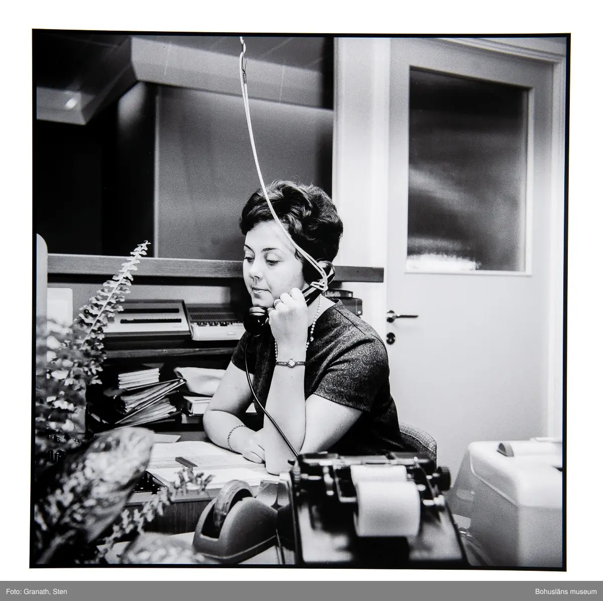 Växeltelefonisten Anita Fransson på Domus sitter vid ett skrivbord och talar i en telefon som hänger i ett snöre från taket.