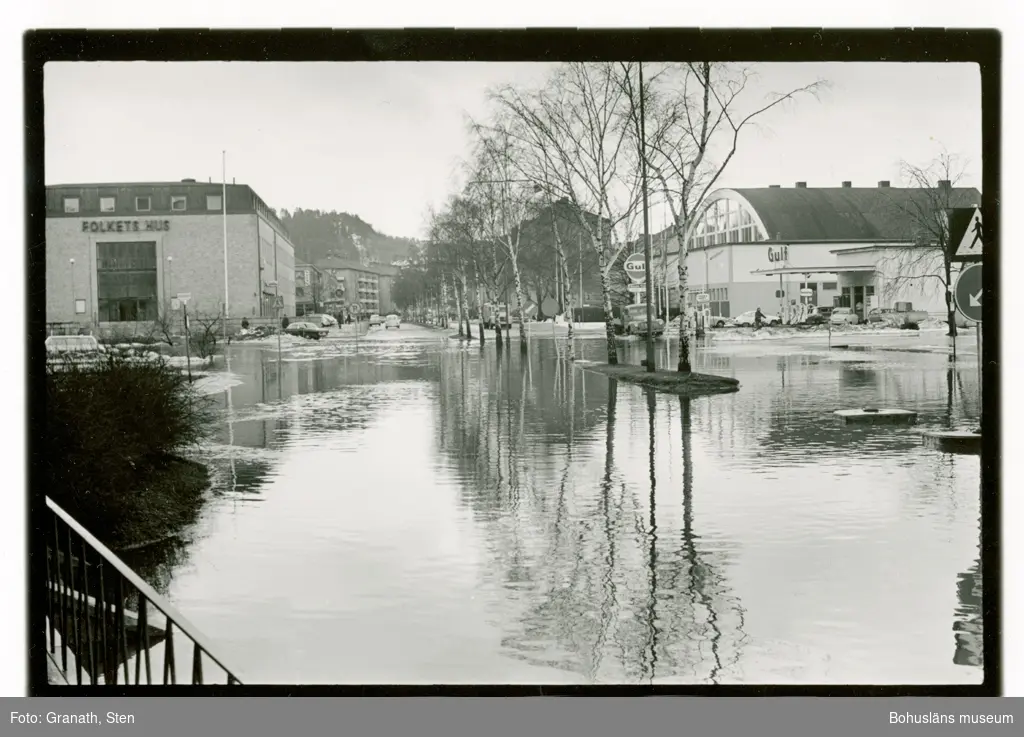 Översvämning på Göteborgsvägen, Uddevalla. I övre halvan av bilden syns Folkets Hus, en Gulf-mack och idrottshallen. Några bilar kör genom vattenmassorna. Det är snövallar längs vägarna och grenarna på träden i allén är kala. I nedre halvan av bilden syns vattenmassorna i vilka  omgivningarna och byggnaderna speglas.