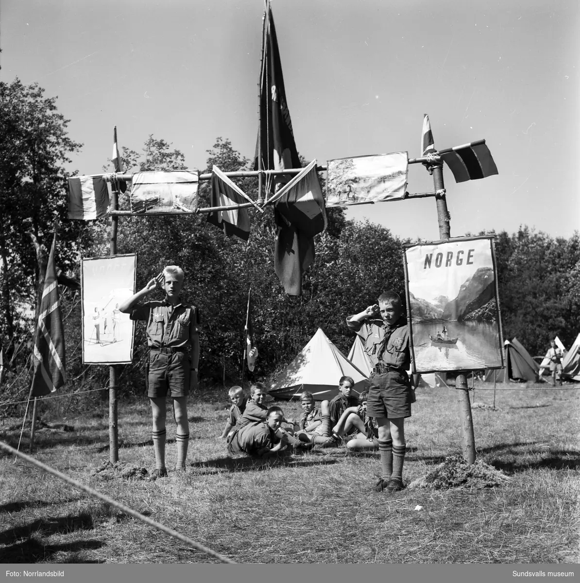 I juli 1959 anordnade Svenska Scoutförbundet sitt stora internationella läger på Rödön utanför Alnö. På första bilden ses lägerledaren Tage Allegård anlända med en grupp pojkscouter för några intensiva veckor på ön. Lägret hade förberetts under ett helt år och dagarna innan hade 70 pojkscouter och ledare hjälpts åt att bygga upp de olika lägerbyarna, som fick namn efter olika fyrar.  SCA hade upplåtit en pråm som låg för ankar i hamnen fylld med de matvaror som krävdes för att mätta de cirka 800 deltagarna.