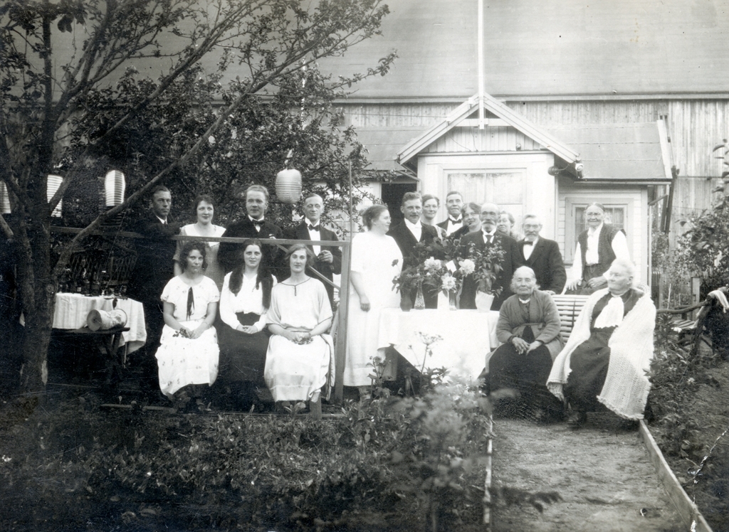 Fotografi taget vid Alfred Träffs förlovningskalas. I trädgården utanför ett litet hus står och sitter ett större sällskap. De är festklädda och från ett träd hänger lyktor.  Alfred är troligen den yngre mannen som står bakom bordet med blomsterbuketter på och hans fästmö tros vara kvinnan till vänster om honom.