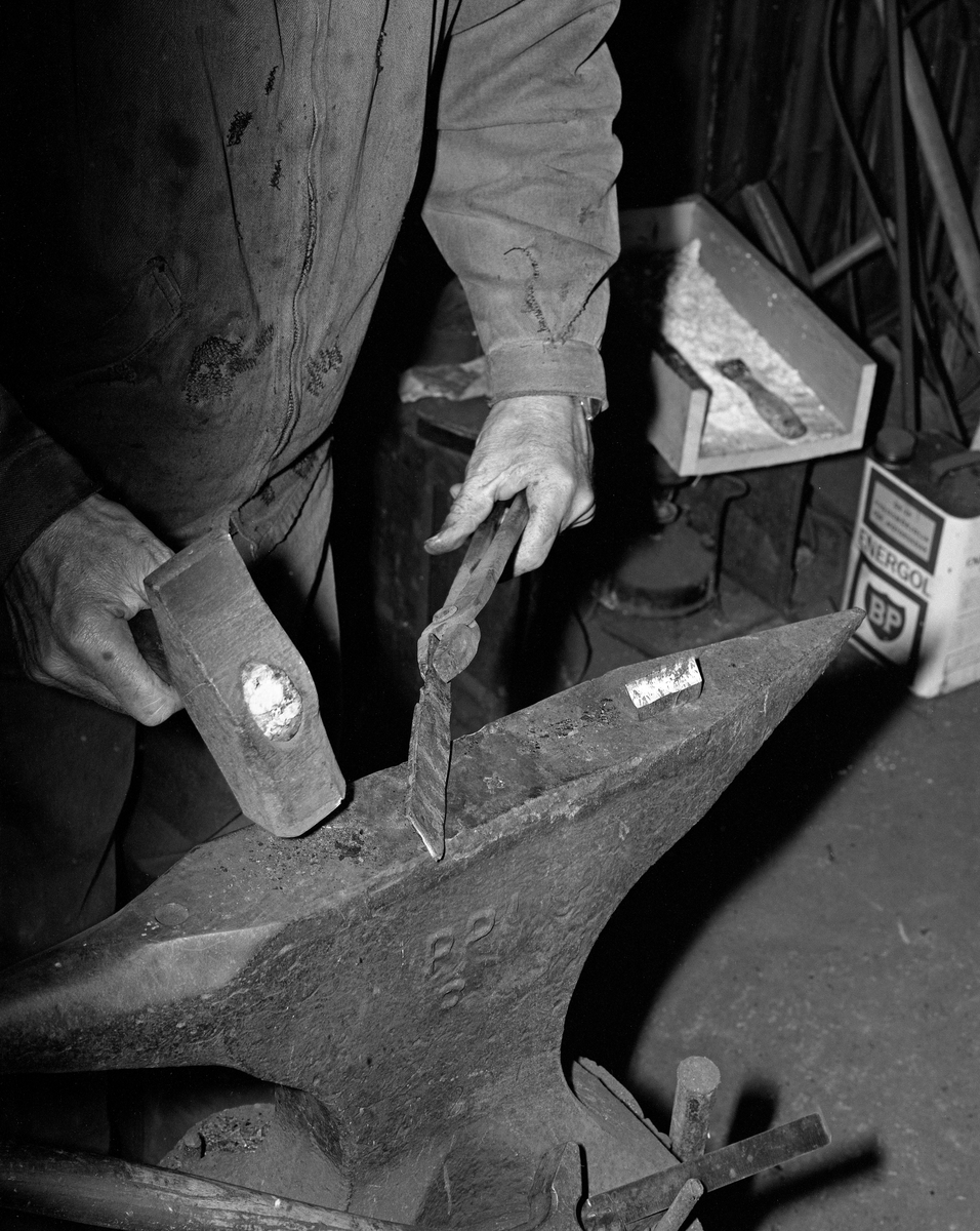 Knivmakeren Even Johannes Blindingsvolden (1908-1985) fra Ringsaker, fotografert i arbeid ved ambolten i smia.  Han holdt et varmt jernstykke i ei tang mens han bearbeidet det med en tung hammer.  Bak den venstre handa til Blindingsvolden ser vi et trebrett med boraks, som ble drysset på kontaktflatene når knivsmeden skulle sveise sammen jern og eggstål i et blademne.  Den på dette tidspunktet 65 år gamle håndverkeren var kledd i dongeriklær mens han arbeidet.  Blindingsvolden eide og bodde på småbruket Rudsland på Rudshøgda.  Her hadde han et et 43 kvadratmeter stort hus ved Prestvegen som inneholdt smie- og verkstedrom.

Mer informasjon om knivsmeden Even Blindingsvolden finnes under fanene «Bildehistorikk» og «Opplysninger».