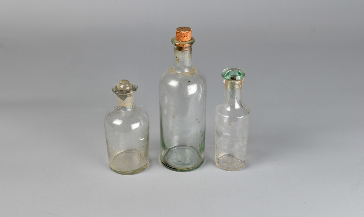 Rette og runde flasker i glass med innsnevring til halsen. Rundt hals. A og C har glasskork, B har kork. A har korken delvis knust på toppen.