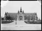 Nordiska museets huvudbyggnad strax före färdigställandet och invigningen 1907. Framför byggnaden står några förbipasserande människor och betraktar byggnadsarbetarna som står på ställningar framför entrén och tornen.