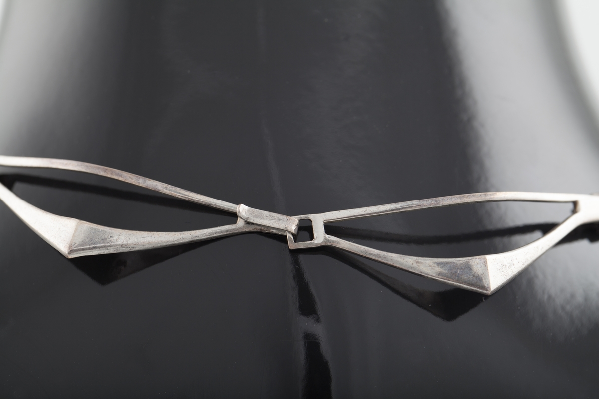 Halssmykke som består av sju ledd som er hektet sammen ved hjelp av små kvadrater. Hvert ledd har form som en halv rombe. "Diagonalen" nærmest halsen er en tynn sølvstreng. "Hjørnet" har en knekk. Fra dette smalner leddet mot yttersiden og ender i en krok. Disse hektes inn i det kvadratiske elementet. De ytterste leddene ender i en krok og et kvadrat som utgjør låsen.