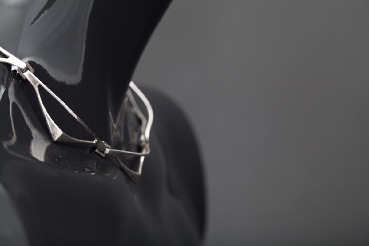 Halssmykke som består av sju ledd som er hektet sammen ved hjelp av små kvadrater. Hvert ledd har form som en halv rombe. "Diagonalen" nærmest halsen er en tynn sølvstreng. "Hjørnet" har en knekk. Fra dette smalner leddet mot yttersiden og ender i en krok. Disse hektes inn i det kvadratiske elementet. De ytterste leddene ender i en krok og et kvadrat som utgjør låsen.