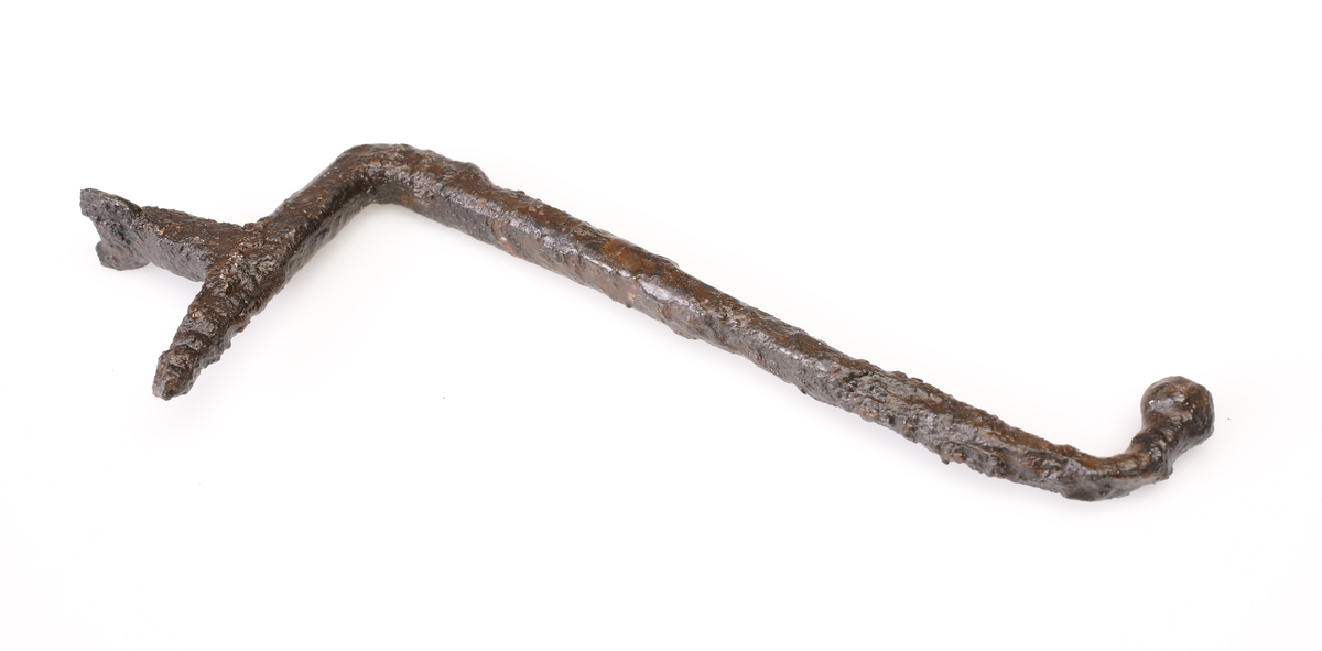 Rund böjd ten som avslutas med en ändknopp i ena änden. Anda änden rätvinkligt böjd och överst gängad och försedd med ett vinkelrätt utskott. Detta utskott är avslutat med en nu ofullständig rund platta med hål. Troligen en avtryckare till ett armborst eller annat handvapen.