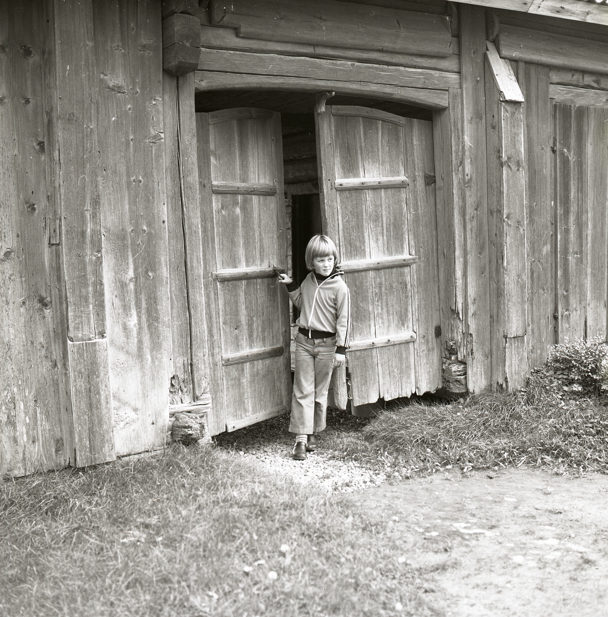 En pojke står i en dörröppning och håller i handtaget till dörren som sitter på en timrad byggnad vid Hembygdsbyn, 4 november 1978.