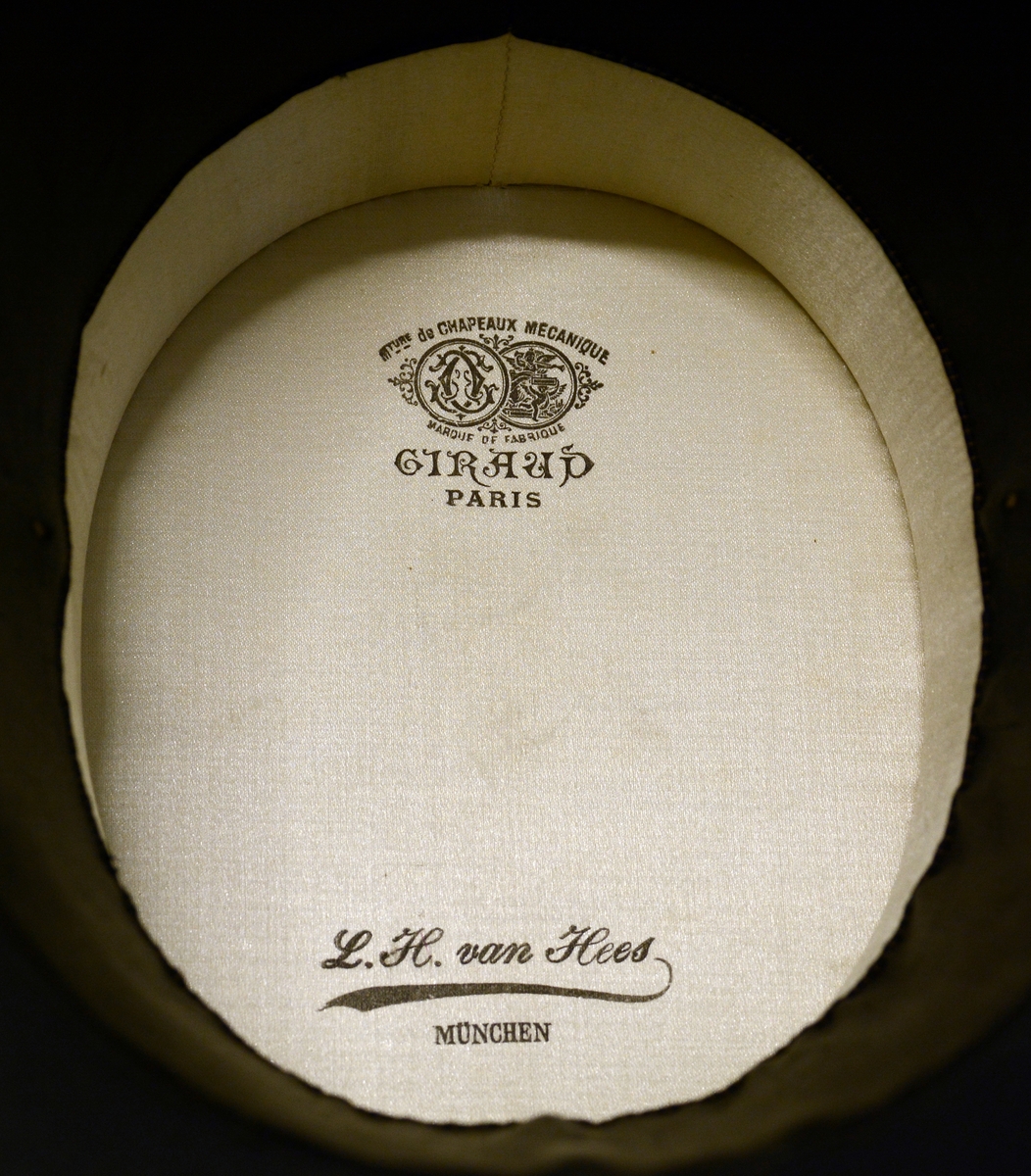 Flosshatt med etui. Sort, sammenleggbar flosshatt eller Chapeaux claque. Silketrukket papp med bånd nederst. Monogram HI med metallbokstaver innvendig. Ovalt etui i brun papp med påskrift i gull: "MANUFACTURE DE PARIS CHAPEAUX MÉCANIQUES PERFECTIONÉS", samt våpenskjold med blant annet skip i midten. Strikk og metallfester på sidene. Kjøpt av firmaet L.H.van Hees, München. Fabrikkert av Giraud, Paris.