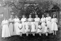 Gruppeportrett av 14 unge kvinner og jenter i lyse kjoler på