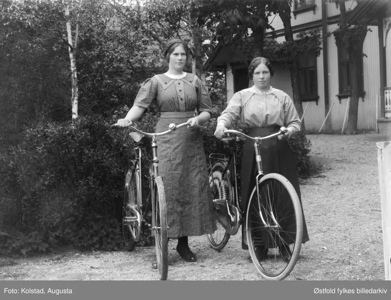 To unge kvinner med sykkel  på tunet utenfor våningshus i Askim ca. 1910 - 1920. Begge kvinnene er ukjente. ØFB.AKO.00022. 