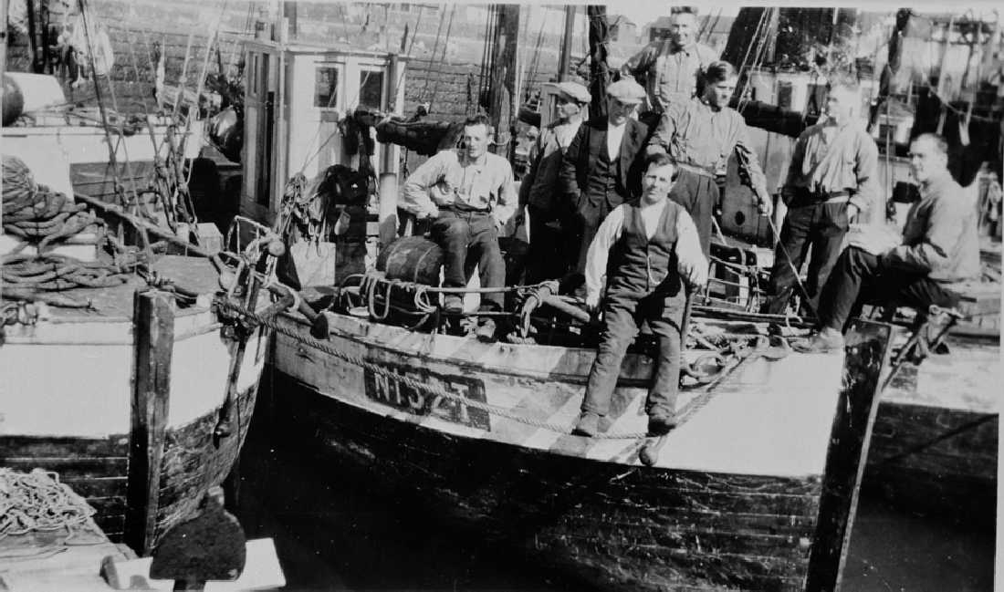 Leirfjord / Vardø, Vardøvågen. 1. pinsedag i vågen. Skipper på skuta er Tore Utnes (sittende på ei tønne). Johan Grønvik (sittende på ripa). Resten er ukjente.