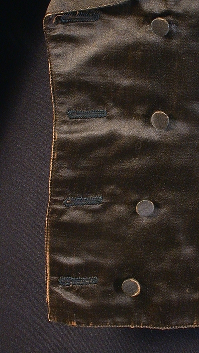 Med framstycke av svart siden, ryggstycke av svart domestik. Sleifar i ryggen med mässingsspänne. Foder av vit lärft. Dubbelknäppt, med 4 stycken svarta knappar i varje rad, på västens innersida en knapp med profilhuvud.