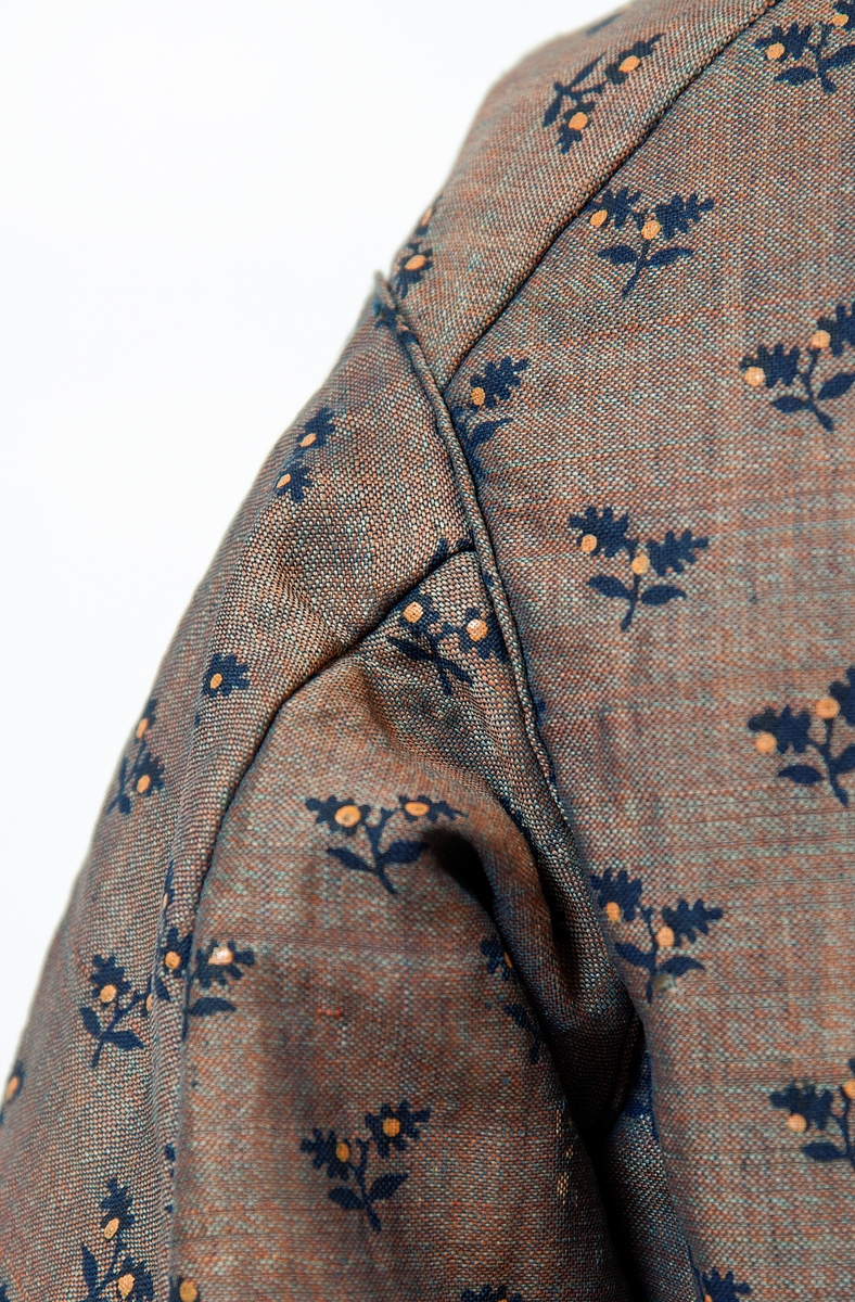 Kjole sammensydd av liv og skjørt med belte i livet med sløyfe i samme stoff. Halvull verkenstoff med trykte blomster. Sydd i Sørum i Akershus ca 1860 og senere brukt på Hamar. Skjørtet har mye håndsøm men vi ser også maskinsøm. Skjørtet er fóret med en skoning av lin i nedre kant. Det er også festet et grønt smalt bånd som slitekant. Overdelen er fóret med ufarget linstoff. Svart fløyelsbånd er festet 1,5 cm fra ermekanten.