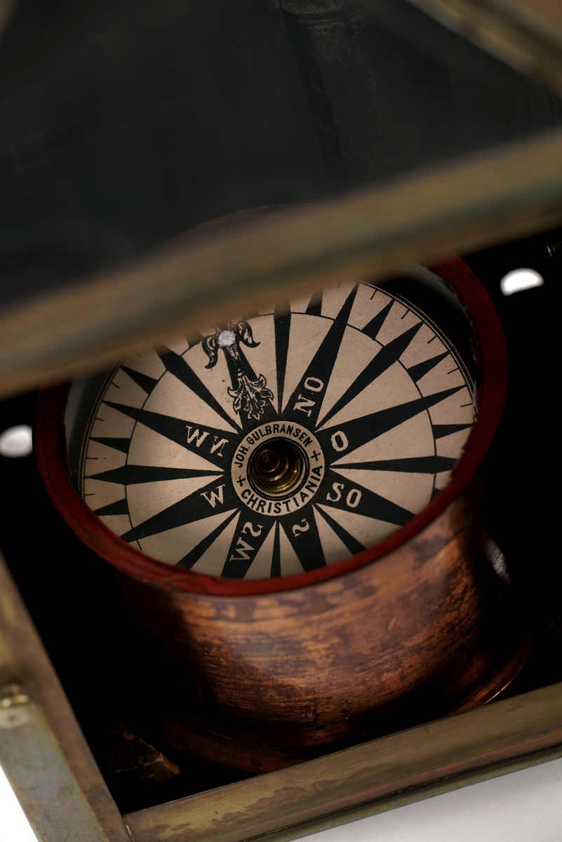 Firkantet kompasshus av metall med dør med vindu av glass. Døren kan låses med krok. Fire forsirede føtter av metall.  Tak som skråner til alle fire sider med vindu på takets forside. Luftavtrekk. Toppen av taket er utstyrt med forsiret deksel med bærering. To støttekroker, èn på hver side. Inne i huset er det to stearinlysholdere med metallfjær-innretning. Mellom disse er det fastmontert et metallrør.
Videre medfølger det to støtteklosser av tre for kompasset. Kompasslokk følger også med.