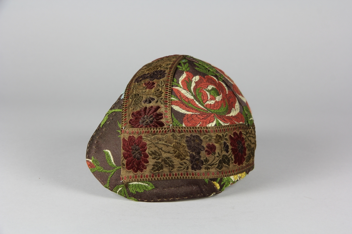 Bindmössa av hårt stärkt textilstomme. Klädd med blommig brokad med brun botten. Runt mössan är ett brett blommigt sidenband fäst med nålar.