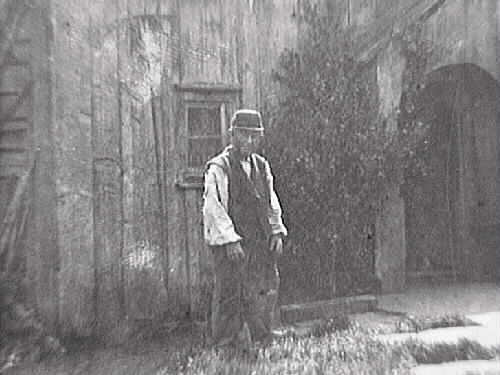 Ryggåsstuga med bislag för ingången, s k skunke (skånke). Framför huset är marken belagd med stenhallar och där står en man i väst och keps. Bilden märkt "Carl Heintz".