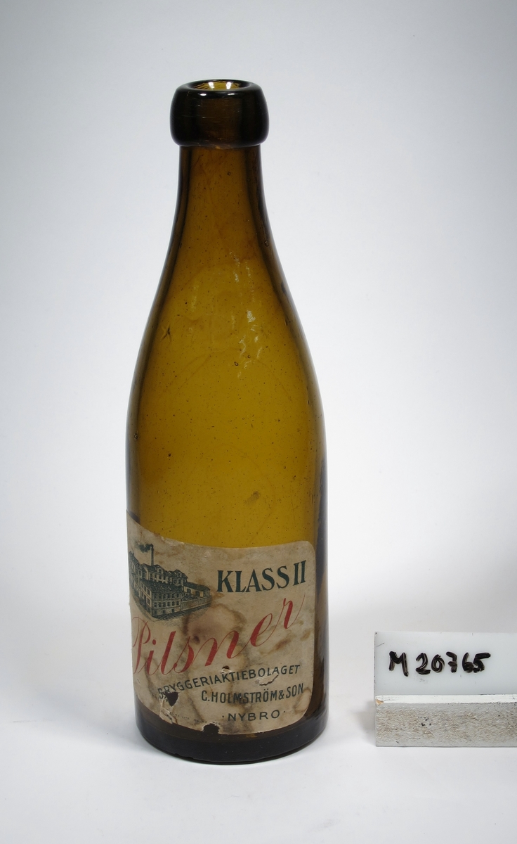Ölflaska i grönt glas. Knoppölsmodell.
Märkt på glaset med ett B omgivet av tre + (det ena ovanför B:et).
Fyrkantig etikett med text: "Klass II Pilsner, Bryggeriaktiebolaget C. Holmström & Son Nybro".
Märkt i botten "1924". 
Inskrivet i huvudkatalogen 1968.