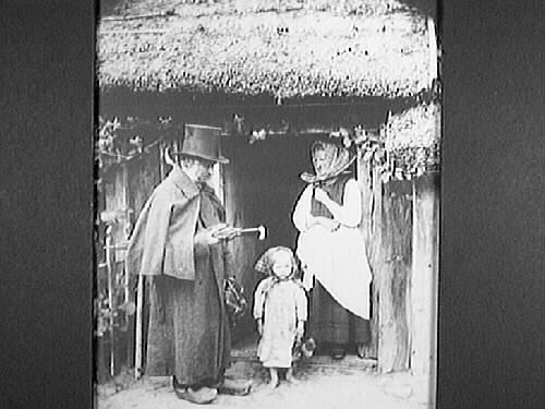 Arrangerad bild, baksidestext: "Hemkomsten från auktionen. Far har varit på fattigauktion anno 1875". Oklart om scenen ska föreställa 1875 eller om fotografiet togs 1875. Man i hög hatt, cape, träskor och paraply med en liten flicka och i dörröppningen står en kvinna. En kaprifol slingrar sig längs husfasaden.