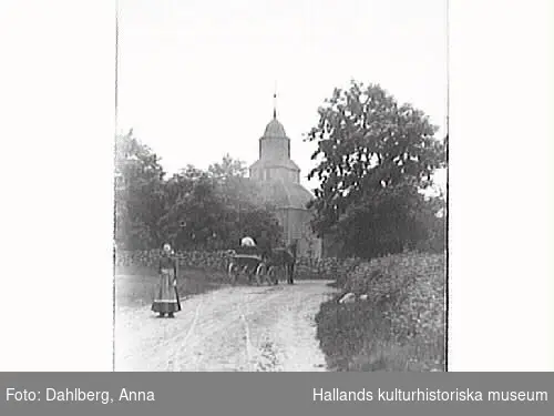 Okome gamla träkyrka. I förgrunden står en kvinna och bakom henne rullar ett hästekipage bortåt på den sandiga vägen.

 Se Information, även tidigare bildnr.