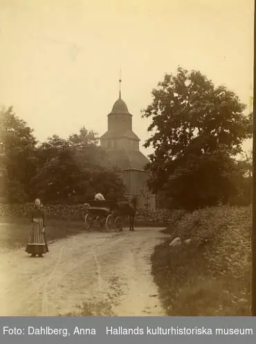 Okome gamla träkyrka. I förgrunden står en kvinna och bakom henne rullar ett hästekipage bortåt på den sandiga vägen.

 Se Information, även tidigare bildnr.