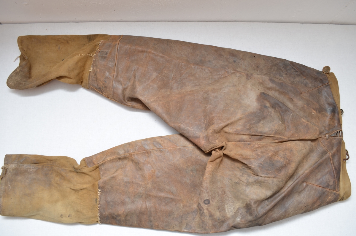 Brun skinnbrok (langbrok). Nedre del av buksebeina er i brunt bomulltøy. (26 cm). (Kan det ha vore kortbrok tidlegare? ) Linning og for i gylfen er i same bomullstøyet.To stikklommer framme  med blått bomullstøy på innsida. Gylf. Open saum midt bak (7 cm), hekta saman med ei skinnreim. Forskjellige knappar.