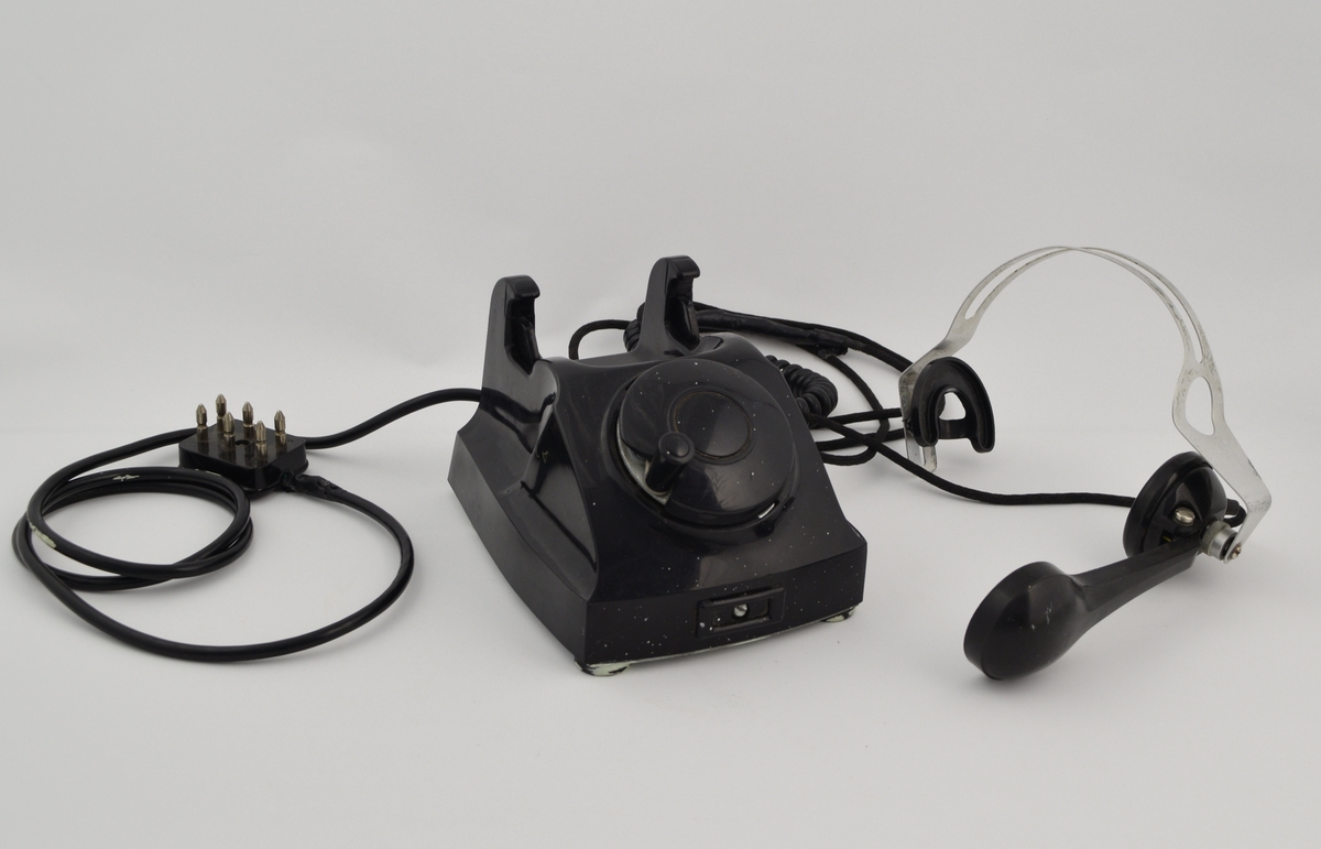 Telefonapparat med telefonrør festet til en hodebøyle i metall med ledning i mellom rør og apparat. Apparatets grunnflate er avlang heksagonformet (sekskantet). Skråner nedover foran der det er plassert en rund nummerskive som er dekket av et rundt deksel med sveiv. Skråner ned bak der gaffelen er plassert, med plass til å plassere hodebøylen med røret. En hodemikrotelefon er koblet til apparatet og har erstattet det originale telefonrøret. Hodemikrotelefonen består av en hodebøyle med høyttaler i venstre øre, og det er der festet en mikrofon fra denne i et ledd som gjør den justerbar.