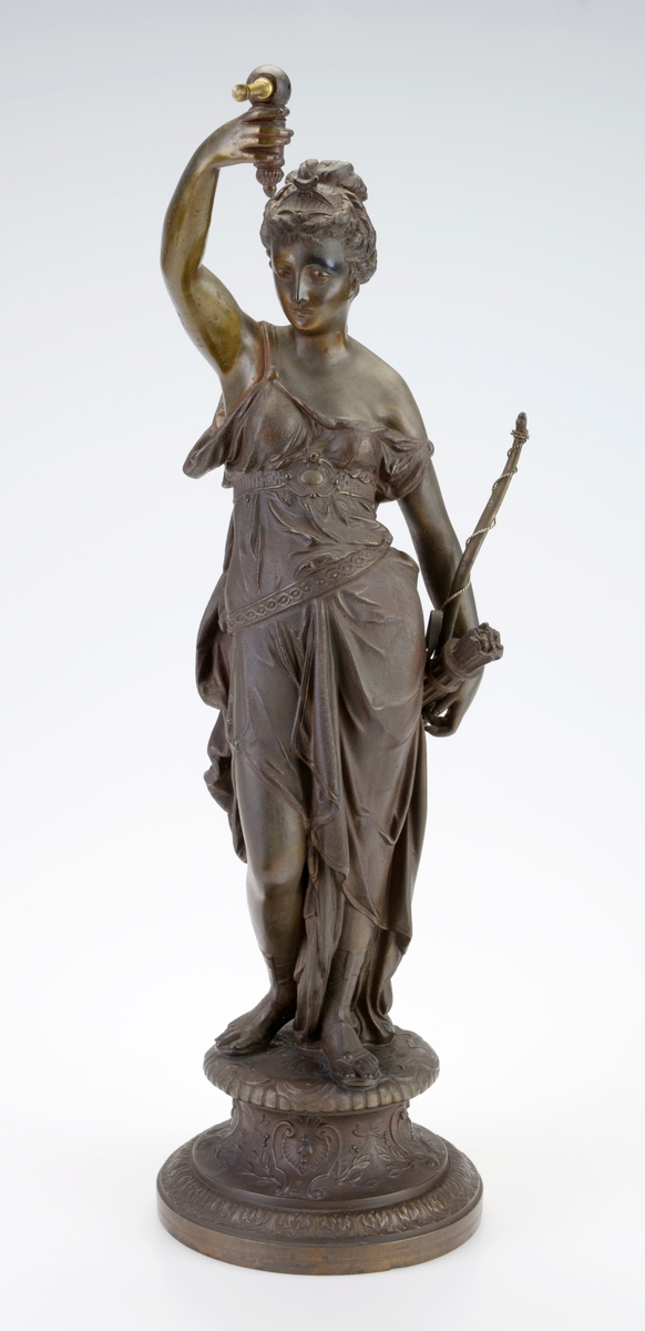 En pendelklokke til å ha stående på et bord, en hylle eller lignende. Den består av en figur/liten statue og en pendel med urverk. Figuren er av støpt metall som er påført maling/lakk slik at det ligner på bronse. Figuren står på en 9,5cm høy sokkel. Det er støpt dekor inn i sokkelen. Figuren forestiller Diana som i romersk mytologi var jaktens gudinne og assosiert med ville dyr, skogen og månen. Hun har en løstsittende kjole med belte og på beina har hun sandaler. Håret er satt opp og i front har hun en kam/liten tiara. Ved den ene hånden har hun bue med sølvfarget metalltråd tvunnet rundt. På denne siden henger det også en kogge med piler. Den andre hånden holder hun over hodet og holder en gjenstand som pendelen skal henges på. Pendelen er laget av forgylt messing og metall. Øverst er det en rund urkasse av blankt metall som er festet i forgylt "messinggrep". Urskiven viser tall fra romertall I - XII. Viserne er laget av svartlakker metall. På urskiven er det informasjon om produsent, se "Påført tekst/merker". På baksiden kan man se urverket som er laget av messing (visst nok et 8-dagers urverk). Fra festet til urkassen henger det to sølvfargede metallstenger med en messingfarget metallstang i midten. Nederst på pendelen er det en utsmykket ramme med et et kvinneportrett i. Dette er støpt og gullforgylt.