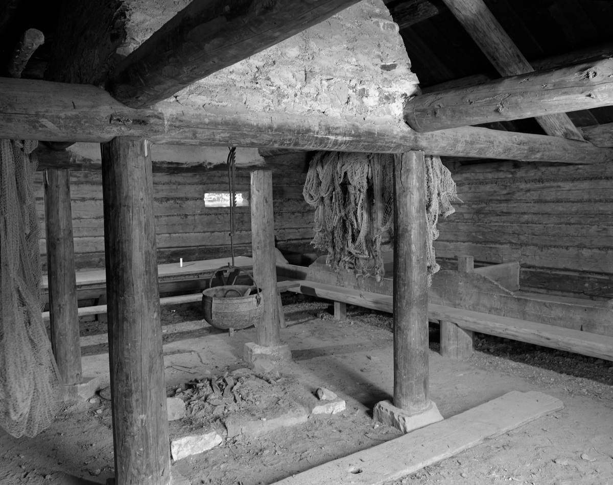 Interiør fra den gamle «Knuvelbua», som opprinnelig stod ved fiskevarpet Knuvelen i Gudbrandsdalslågen, en grusbanke nokså rett vest garden Storhove i Fåberg.  Bygningen er seinere demontert og flyttet til friluftsmuseet på Maihaugen i Lillehammer by, der den er plassert ved «Breisjøen».  Har framstår Knuvelbua som en laftet bygning med jordgolv.  Interiøret er enkelt.  Det besto av faste senger med en halvkløvningsbenk langs den ene veggen og et langbord langs den andre.  Midt i rommet var det et åpent ildsted på en lav elvpall av tilhogd stein.   Omkring dette sto det fire stolper, som støtter for krysningspunktene mellom tverrgående beiter og langsgående slinder i rommets raftsone.  Oppå disse var det murt et pipeløp.  Da dette fotografiet ble tatt hang det ei skjerding med ei støpt jerngryte over ildstedet og det hang nøter ved siden av ildstedet.  På veggen i bakgrunnen skimtes en smal lysåpning.  Bygningen ble opprinnelig brukt som kvilested for menn som deltok i det store sesongfisket etter lågåsild (Coregonus albula) i den nedre delen av Lågen.