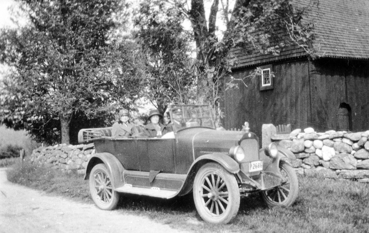 Anna Norlander på biltur med kvinna och en manlig chaufför sitter i en personbil utanför Stavkyrkan i Hedared.
Bilen, med registreringsnummer P2686,  är en Star 1924 och står parkerad framför en stenmur.
