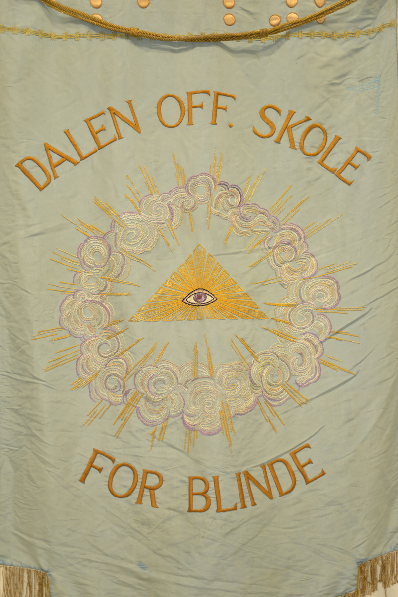 Rektangulær fane. Dobbeltsidig duk med tekst og broderier på begge sider. Fanen er festet med løkker på en lakkert trestang. I endene av denne stanga er det dekorasjon i form av stjerner. Stjernene er laget i tre og malt med gullfarge. Det henger to snorer på hver ende av stanga; den ene med dusk i enden, den andre har en løkke i enden. 

Fanen er tosidig og er laget av silke. Den ene siden har blå bakgrunnsfarge, punkter (punktskrift), tekst og påbrodert motiv (skyer, solstråler, stjerne med øye). Den andre siden har lys gul farge, en bord, tekst og dato. Nederst på faneduken henger det frynser av gullfarget tekstil. 

Sammen med fanen er det en lang stang som består av to deler som festes i hverandre. Fane med stang festes i denne lange stanga ved hjelp av en metallkrok. Øverst i den lengste stanga er det en gullfarget stjerne av tre.