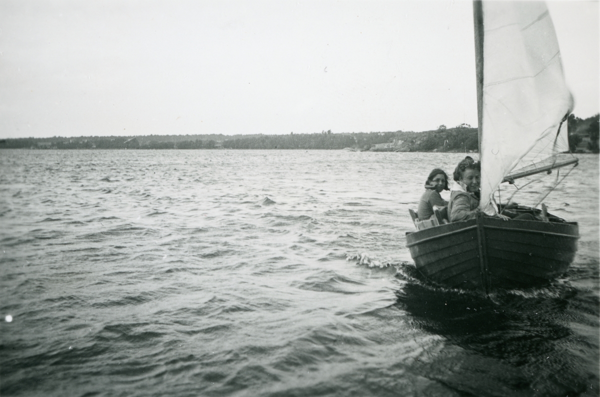 Två kvinnor seglar en mindre segelbåt. Landskap i bakgrunden.