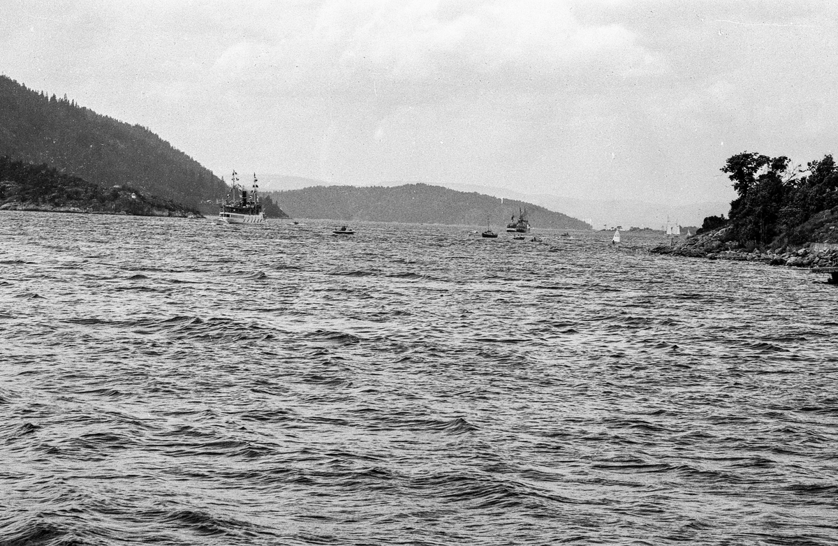 Drøbaksfjorden, sundet ved Oscarsborg. Tre menn på brygga ser på båtene med kikkert. Sandtaket og småhus på Hurum.
Fotograf: ØB Rønning