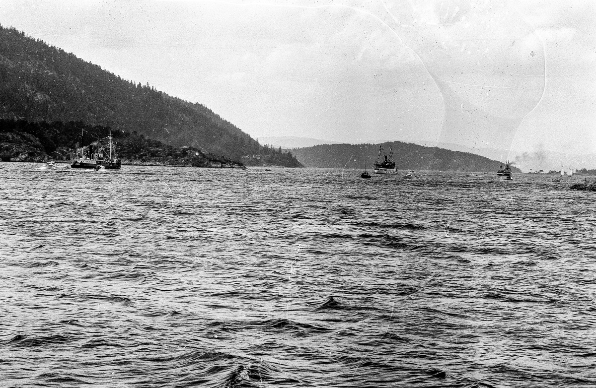 Drøbaksfjorden, sundet ved Oscarsborg. Tre menn på brygga ser på båtene med kikkert. Sandtaket og småhus på Hurum.
Fotograf: ØB Rønning