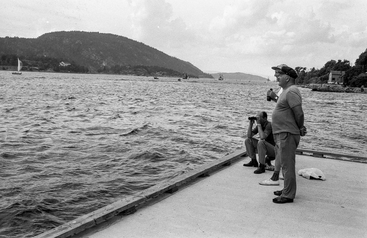Drøbaksfjorden, sundet ved Oscarsborg. Tre menn på brygga ser på båtene med kikkert. Sandtaket og småhus på Hurum.
Fotograf: ØB Rønning