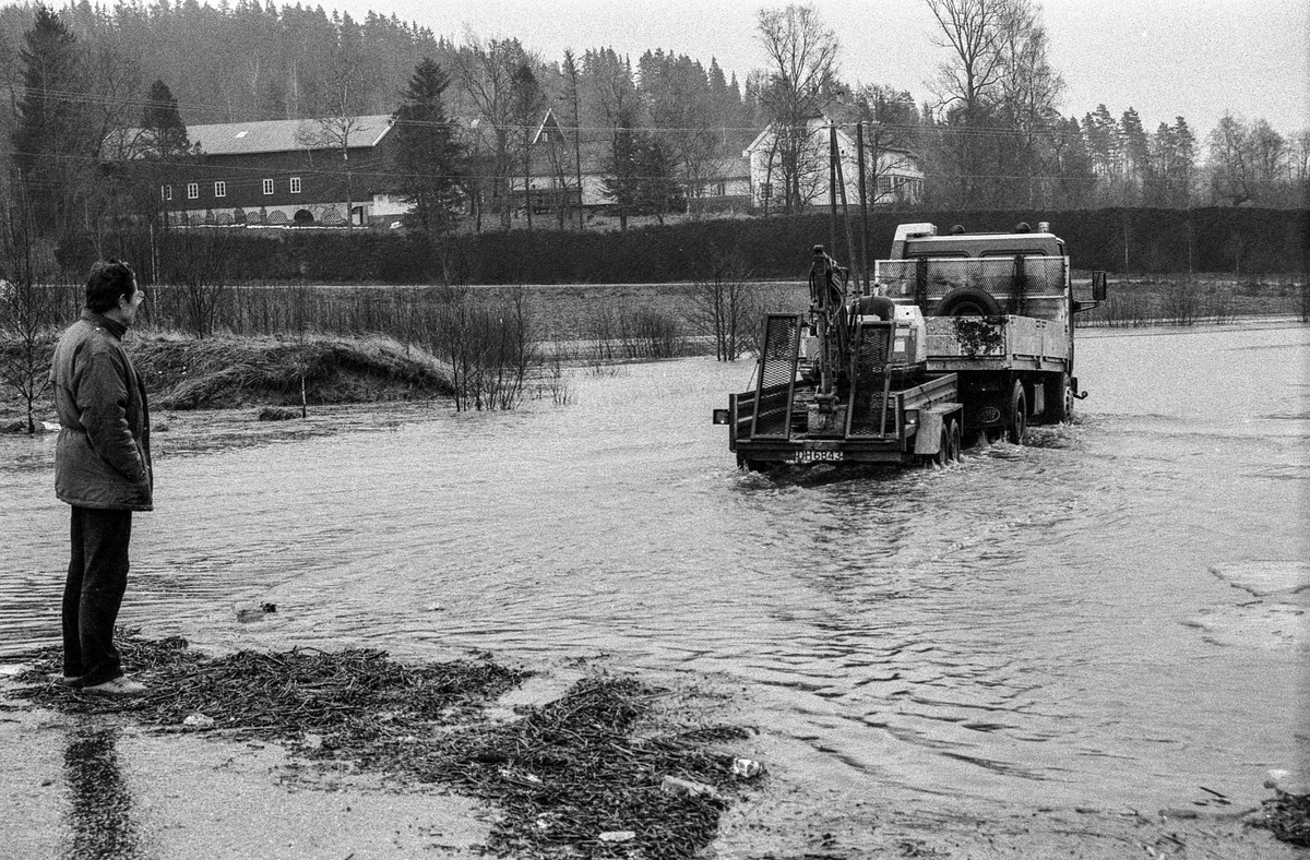Flomskader på veiene, Riksvei 120, Ytre Enebakk. Vannet flommer over veier og jorder, biler kjører så vannspruten står.