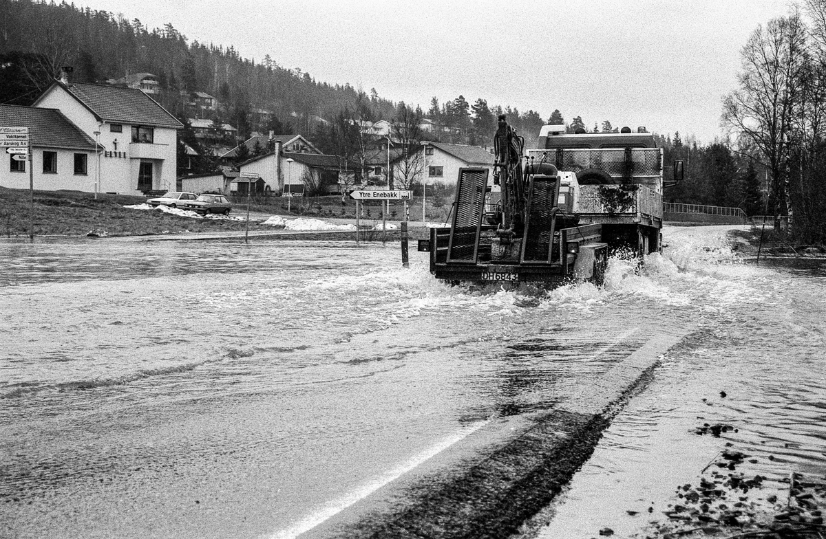 Flomskader på veiene, Riksvei 120, Ytre Enebakk. Vannet flommer over veier og jorder, biler kjører så vannspruten står.