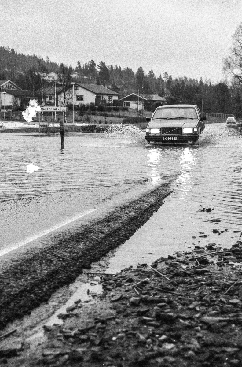 Flomskader på veiene, Riksvei 120, Ytre Enebakk. Vannet flommer over veier og jorder, biler kjører så vannspruten står.