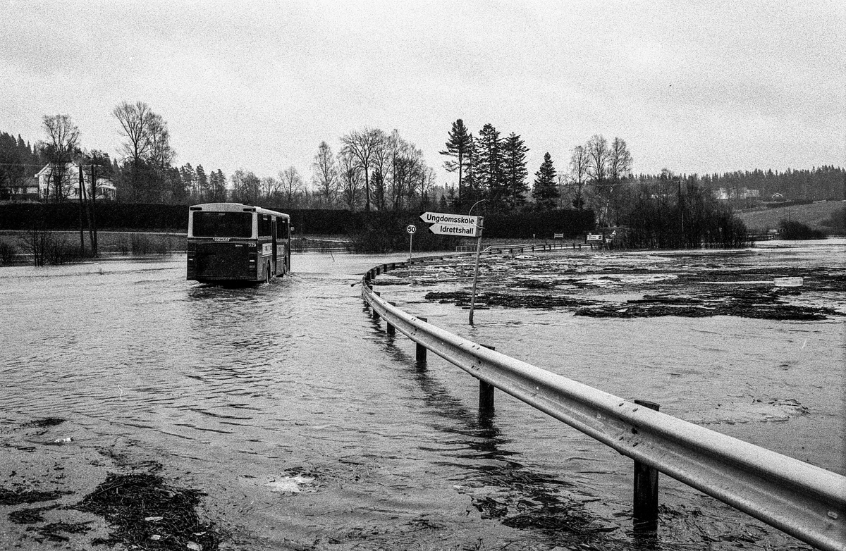 Flomskader på veiene, Riksvei 120, Ytre Enebakk. Vannet flommer over veier og jorder, biler kjører så vannspruten står.