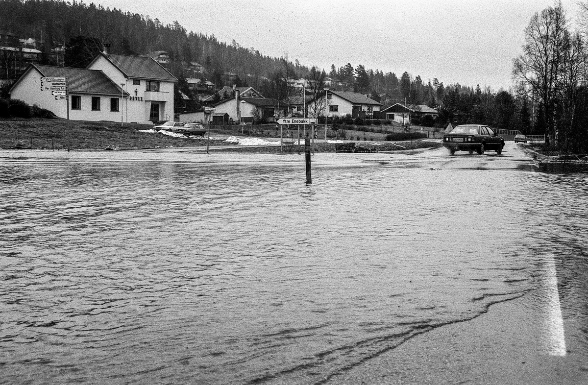 Flomskader på veiene, Riksvei 120, Ytre Enebakk. Vannet flommer over veier og jorder, biler kjører så vannspruten står.