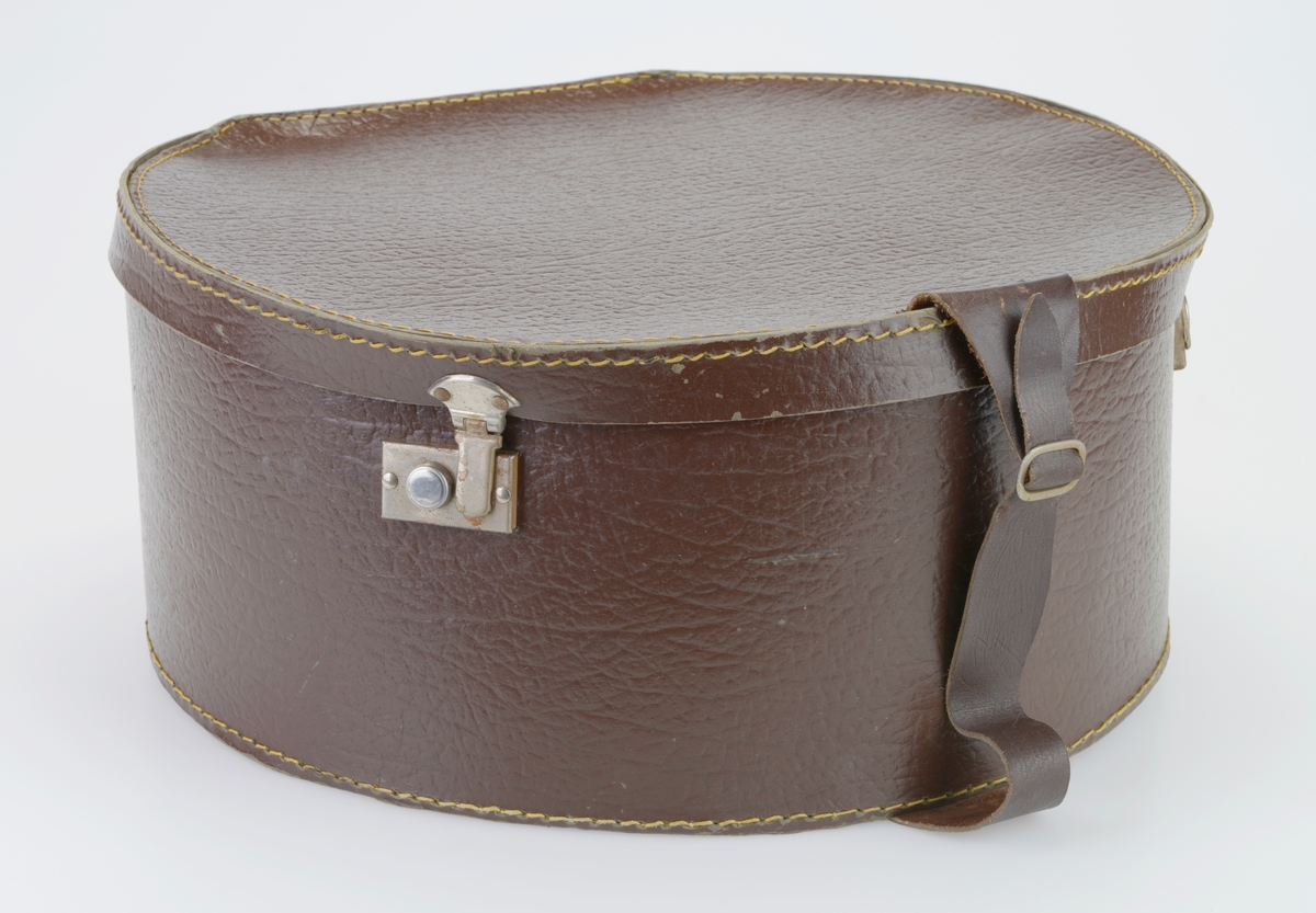 En hatteeske som i all hovedsak er rund men med avskåret bakende. Lokk og eske er laget av preget papp som er grå på innsiden og brun på utsiden. De ulike delene er sydd sammen av kraftig gul tråd og forsterket med brunmalte nagler. I front er det to koffertlåser av metall. I front er det tilskårede lærreimer med en enkel spenne av metall. Lærreimene er festet med brunmalte nagler. På baksiden er det to hengsler av metall som fester lokket til esken.