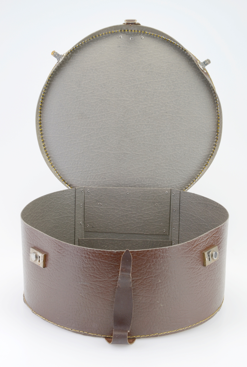 En hatteeske som i all hovedsak er rund men med avskåret bakende. Lokk og eske er laget av preget papp som er grå på innsiden og brun på utsiden. De ulike delene er sydd sammen av kraftig gul tråd og forsterket med brunmalte nagler. I front er det to koffertlåser av metall. I front er det tilskårede lærreimer med en enkel spenne av metall. Lærreimene er festet med brunmalte nagler. På baksiden er det to hengsler av metall som fester lokket til esken.