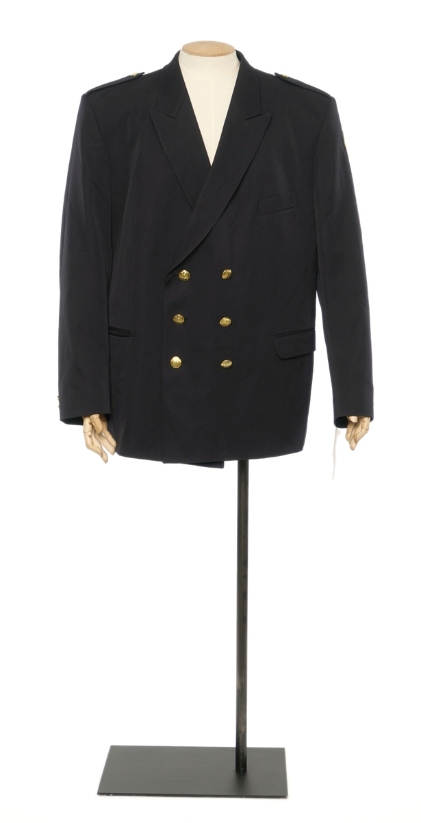 Mørkblå jakke enkelspent med tre riksløveknapper. Epauletter uten distinktsjoner. Tekstil marke på venstre arm, riksløve med hvit øks omgitt av eikeblader over TOLL. Hempe i nakke. Kypert stoff, blanding av polyester og ull. Jakke er foda med antatt syntetisk stoff, mørkblå.
Bukser av samme stoff.