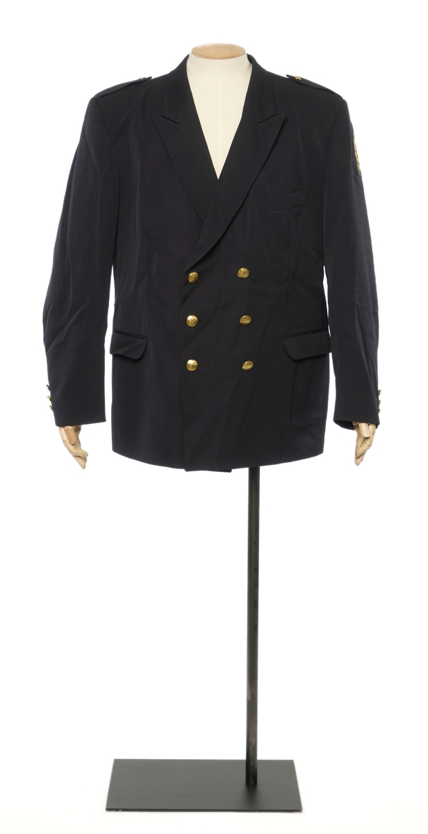 Mørkblå jakke enkelspent med tre riksløveknapper. Epauletter uten distinktsjoner. Tekstil marke på venstre arm, riksløve med hvit øks omgitt av eikeblader over TOLL. Hempe i nakke. Kypert stoff, blanding av polyester og ull. Jakke er foda med antatt syntetisk stoff, sort.
Bukser av samme stoff.