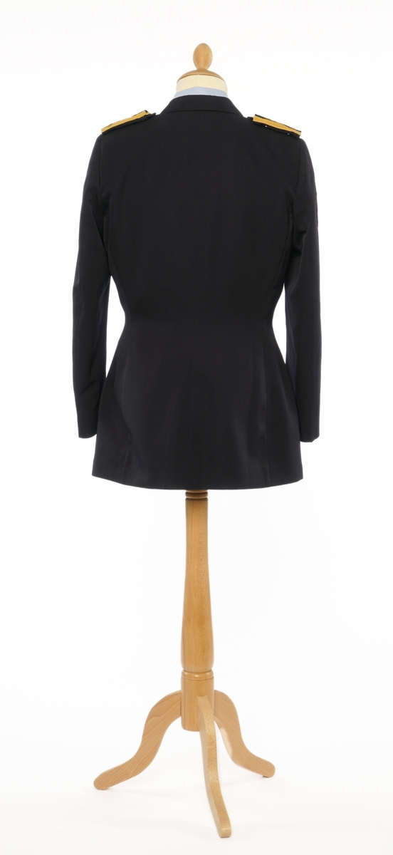 Mørkblå dame jakke enkelspent med fire riksløveknapper. Epauletter med merkurstav, krone, og to kvist eikblader (fire blader totalt) mellom to gullbånd. Tekstil marke på begge armer, krone over riksløve med hvit øks i rød skjold omgitt av eikeblader over TOLL/CUSTOMS. Hempe i nakke. Kypert stoff, blanding av polyester og ull. Jakke er foda med antatt syntetisk stoff, sort.
Hvit hatt med sort visir. Metall distinkjons skilt av krone over riksløve med søkvferget øks i rød skjold omgitt av eikeblader. Gullfletting over visir, og eikeblader på visir.
Lys blå dame skjorte med matchende distinktsjoner og tekstil merker på armene.