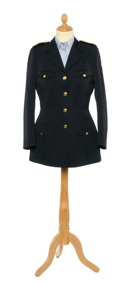 Mørkblå dame jakke enkelspent med fire riksløveknapper. Epauletter med merkurstav, krone, og to kvist eikblader (fire blader totalt) mellom to gullbånd. Tekstil marke på begge armer, krone over riksløve med hvit øks i rød skjold omgitt av eikeblader over TOLL/CUSTOMS. Hempe i nakke. Kypert stoff, blanding av polyester og ull. Jakke er foda med antatt syntetisk stoff, sort.
Hvit hatt med sort visir. Metall distinkjons skilt av krone over riksløve med søkvferget øks i rød skjold omgitt av eikeblader. Gullfletting over visir, og eikeblader på visir.
Lys blå dame skjorte med matchende distinktsjoner og tekstil merker på armene.