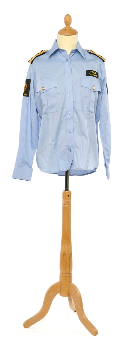 Lys blå langermet skjorte av et fintvevd stoff. Epauletter med tilsatt distinktsjon: krone over merkurstav over en kvist eikeblader (fire blader totalt), med gull bånd på begge sider. Tekstil merke av krone over riksløve med hvit øks i rødt skjold  omgitt av eikeblader over TOLL/CUSTOMS på begge armer.